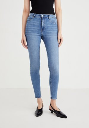 Personne portant un jean skinny bleu clair, un top noir sans manches et des talons bas noirs à bout pointu et bride arrière, debout sur un sol blanc.