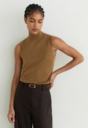 Vrouw met vlechten, gekleed in een mouwloos bruin coltruitje en een zwarte broek met een zwarte riem, staat tegen een witte muur.