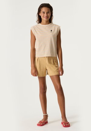 Junge Frau trägt beige gestreiftes ärmelloses Oberteil, hellbraune Shorts, rote Flip-Flops und ein Muschel-Fußkettchen vor einfarbigem Hintergrund.