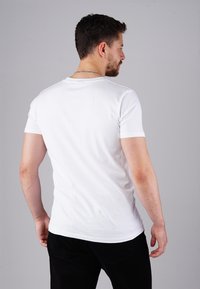 Mann mit kurzem dunklem Haar und Bart, der ein weißes Kurzarmhemd und schwarze Hosen trägt, steht mit dem Rücken zur Kamera vor einem grauen Hintergrund.