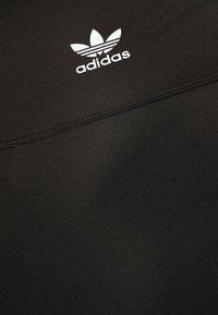 Melnā sporta auduma ar gludu faktūru, kurā redzams baltais Adidas logotips un trīs horizontālās svītras. Detalizējumu veido plakani šuvumi.