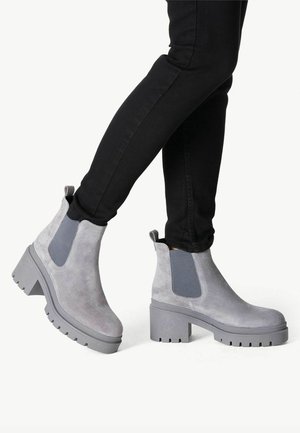 Bottes de cheville en suède gris avec des panneaux élastiques sur les côtés, un large talon block, une semelle texturée et une languette à l'arrière. Portées avec un jean skinny noir.
