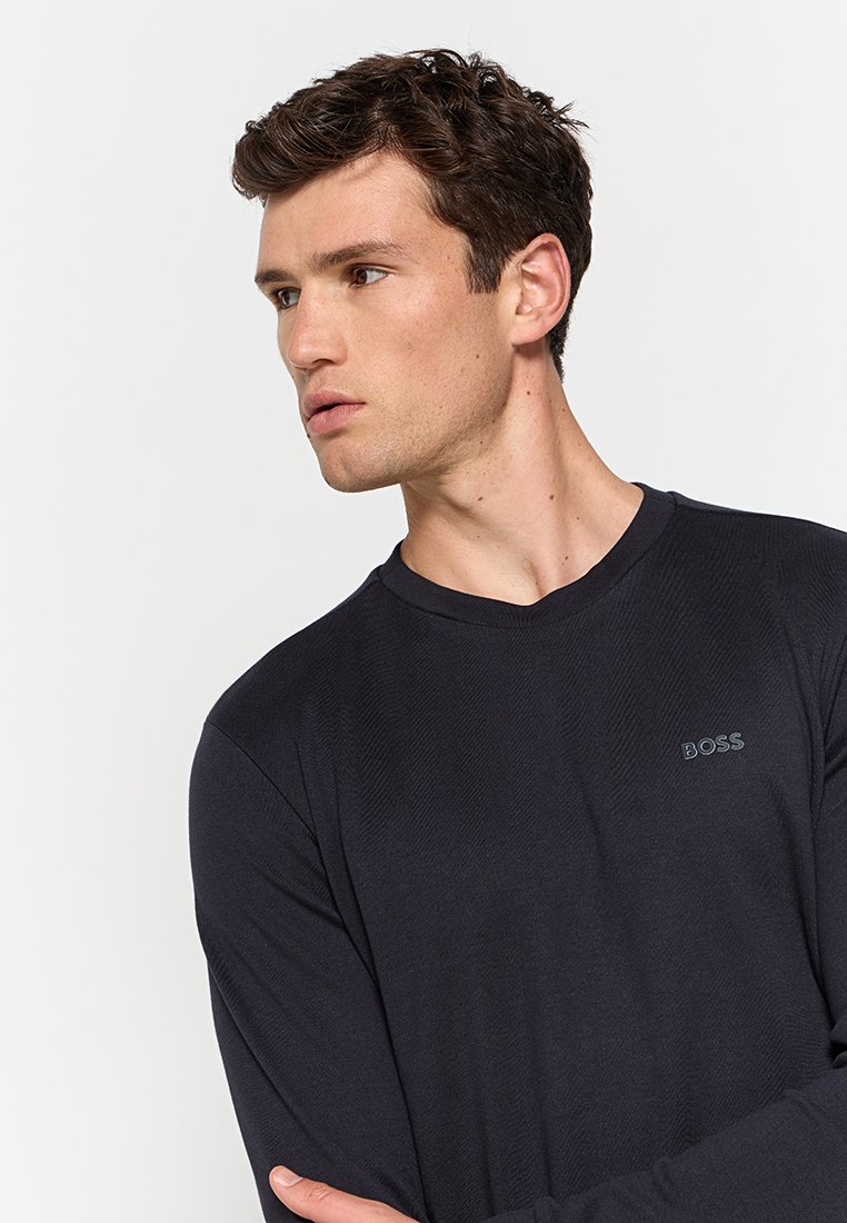 Chemise noire à manches longues avec un col rond, arborant un logo discret sur la poitrine et un tissu texturé. Design ajusté.