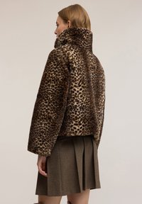 Kobieta w brązowej kurtce z imitacji futra w panterkę z szerokimi rękawami oraz krótkiej brązowej plisowanej spódnicy, widziana od tyłu.