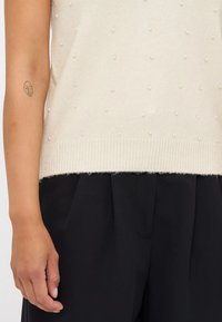 Gebreide trui in crème met textuur van bobbeldetails; zachte stof; geribbelde zoom. Gecombineerd met zwarte loszittende broek, waarbij een deel van een arm zichtbaar is.