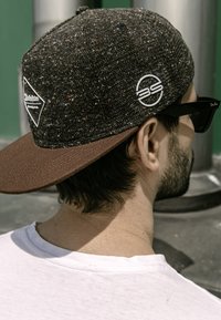Brown-brimmed cap with a textured dark fabric panel, featuring white logos and a flat visor, worn alongside black sunglasses.