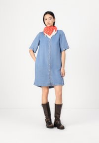Denim ruokopuku, jossa on napit, lyhyet käännettävät hihat ja hieman A-linjan muoto. Yhdistetty polvipitkiin ruskeista nahkasaappaisiin ja punaiseen kuviolliseen huiviin.