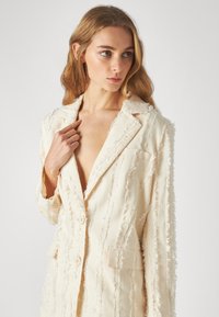 Blazer ajustado en color crema con un diseño texturizado y deshilachado, dos botones y un bolsillo en el pecho, hecho de una tela ligera.