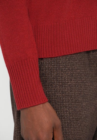 Rode gebreide trui met geribde manchetten, gecombineerd met bruine gebreide broek met een klein houndstoothpatroon. Close-up van de mouw en hand.