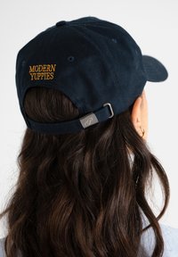 Tummansininen puuvillapipo, jossa on keltainen brodeeraus "MODERN YUPPIES", takana säädettävä hihna metalliosalla.