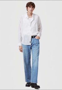 Camisa branca às riscas, com botões, corte solto, um bolso no peito; jeans azuis claros de perna reta; sapatos pretos; fundo neutro.