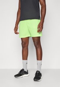 Mann in neongrünen Sportshorts, schwarzen Laufschuhen, grauen Kniestrümpfen und einem dunkelgrauen T-Shirt steht vor einem schlichten Hintergrund.