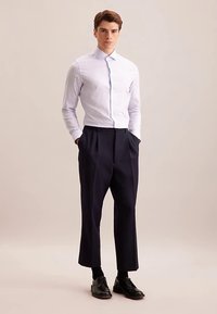 Wit overhemd met lichtblauwe strepen, gecombineerd met donkerblauwe plissébroek. Model staat met handen in de zakken, draagt zwarte schoenen.