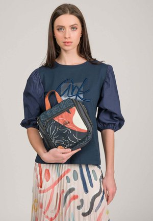 Donna con capelli castani lunghi indossa un top blu navy con maniche a sbuffo e una gonna con stampa astratta colorata, tiene una borsa quadrata a fantasia multicolore.