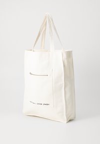 Canvas tote bag in off-white, featuring long handles, a front zip pocket, and printed text near the bottom edge. Simple design, durable material.