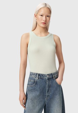 Ung kvinde med langt blondt hår iført en ærmeløs lysegrøn ribbet top og højtaljede blå jeans med den ene hånd i lommen.