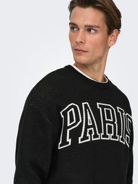 Black sweater with oversized "PARIS" text in white. Features a round neck, textured fabric, and ribbed detailing at the collar and cuffs.