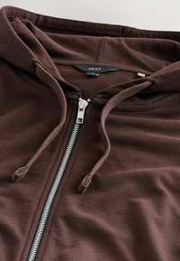 Brauner Reißverschluss-Hoodie aus weichem Stoff, mit silbernem Reißverschluss und Kordeln sowie einem strukturierten Innenfutter, sichtbarer Markenlabel.