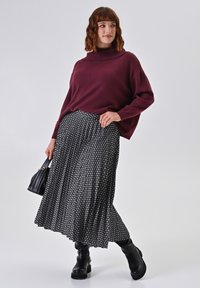 Burgunderfarbenes kurzgeschnittenes Sweater mit hohem Kragen, kombiniert mit einem schwarzen Plisseerock mit einem weißen geometrischen Muster. Getragen mit schwarzen klobigen Stiefeln.