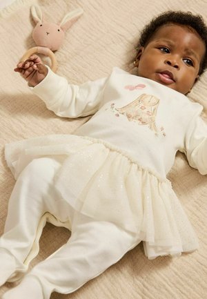 Cremfarbener Overall mit einem glitzernden Tüllrock, der ein Hasenmotiv zeigt. Aus weichem Stoff, mit einer Hand, die ein Holzringspielzeug auf einer strukturierten Decke hält.