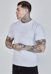 T-shirt grigia a maniche corte con logo ricamato "SIKSILK" sul petto. Il modello ha braccia tatuate e indossa jeans scuri. Tessuto morbido.