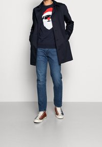 Marinblå kappa över en marinblå tröja med en vit och röd juldamsansikte-design, parat med blå jeans och sneakers med bruna accenter.