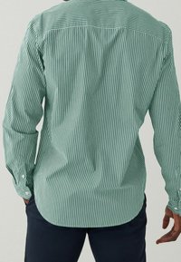 Zielono-pasywana koszula z długim rękawem, z guzikami przy mankietach i zaokrąglonym dołem. Posiada pionowe białe paski na teksturowanym tle.