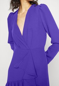 Lila långärmad klänning med djup V-ringning, volangdetaljer på kjolen och ihopdragna axlar, tillverkad av ett mjukt tyg.