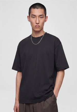 Jeune homme aux cheveux courts portant un t-shirt noir ample, un pantalon marron et un collier chaîne en argent, debout avec une main dans la poche.