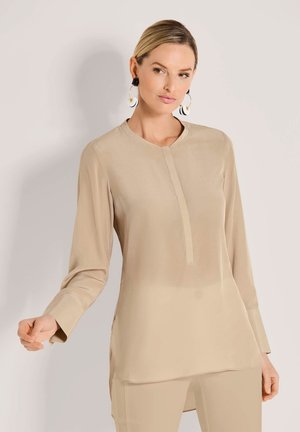 Beige blouse met lange mouwen, een ronde halslijn en een lichte sluiting aan de voorkant. Zacht, glad materiaal met zijsplitten; gedragen met bijpassende broek.
