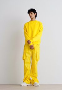 Felpa gialla e pantaloni cargo realizzati in un tessuto morbido, con una vestibilità rilassata e tasche laterali visibili, abbinati a scarpe bianche.