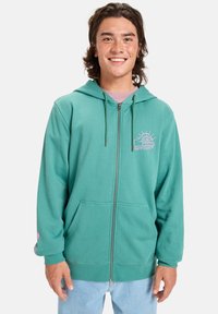 Tyrkysová mikina s kapucňou na zips z mäkkého materiálu, s prednými vreckami, sťahovacou kapucňou a ružovým grafickým logom na hrudi a na rukáve.