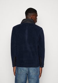 Veste bleu marine avec une texture lisse, dotée d'un col noir et d'accents en tissu noir sur les manches. Vue arrière, design simple.