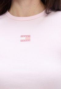 Kerged roosa ribaga T-särk, millel on rinnal väike roosa ja valge "TOMMY JEANS" logo patch. Materjal näib olevat pehme ja keha lähedal.