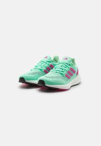 Pár mátově zelených sportovních bot s růžovými pruhy a akcenty, bílé mezipodešve a černé podešve, umístěné vedle sebe na bílém pozadí.