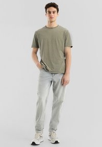 Olijfgroen katoenen T-shirt met korte mouwen, ronde hals en een ontspannen pasvorm; gecombineerd met lichtblauwe jeans en grijze sneakers.