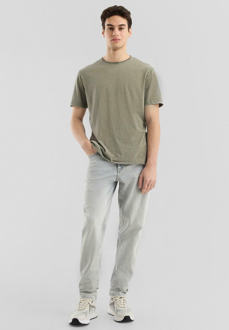 Olivgrünes Baumwoll-T-Shirt mit kurzen Ärmeln, Rundhalsausschnitt und lässiger Passform; kombiniert mit hellblauen Jeans und grauen Turnschuhen.