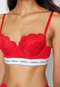 Reggiseno in pizzo rosso e tessuto con coppe imbottite, spalline regolabili e una fascia bianca con il marchio "GUESS" in lettere nere. Bordo ondulato.