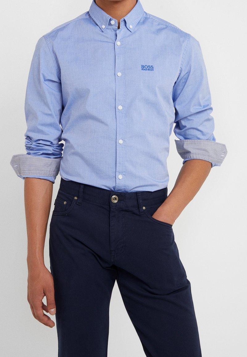 Světlomodrá košile s knoflíky a límečkem, s malým modrým logem "BOSS". Nošena s tmavě námořnickými kalhotami, s ohrnutými pruhovanými manžetami.