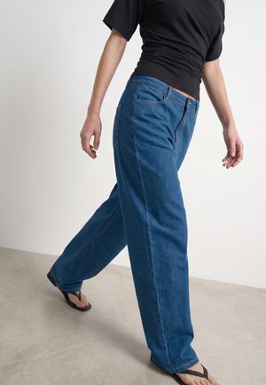 Persoon die blauwe wijde jeans draagt, een zwart T-shirt met korte mouwen en zwarte slippers, lopend op een lichte vloer met een effen witte achtergrond.