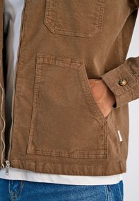 Brun kordjacka med dragkedja, två framfickor och detaljer med knäppning vid ärmarna; bärs över en vit skjorta och blå jeans.