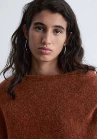 Rust-colored wool sweater with a loose fit and wide neckline. Features long, dark hair and large silver hoop earrings.
