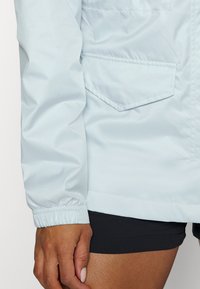 Vänster arm i ljusblå jacka med elastiska manschetter och ficka med lock, hängande bredvid en person med ljusbrun hud som bär svarta shorts.