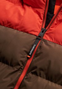 Wattierte Jacke mit einem Farbschnitt-Design, das orangefarbene und braune Abschnitte aufweist, einem auffälligen Reißverschluss und einem sichtbaren Markenetikett.