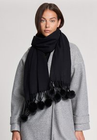 Czarny szalik wykonany z miękkiego materiału, z frędzlami na dole ozdobionymi pomponami. Noszony na jasnoszarej odzieży.