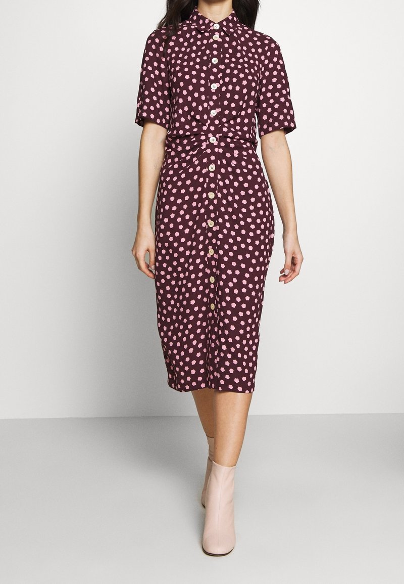 Bordeauxfarbene Kleid mit kurzen Ärmeln, pinken Polka-Dots, Knopfleiste und gebundenem Taillengürtel. Kombiniert mit hellpinken Ankle Boots. Glatte Stofftextur.