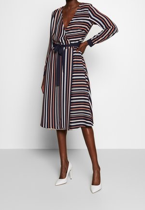 Femme portant une robe portefeuille midi à rayures bleu marine, blanche et pêche avec des manches longues et des talons hauts blancs, debout devant un fond uni.