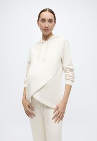 Μπεζ εγκυμονούν φούτερ με σχεδιασμό crossover, μακριά μανίκια και κουκούλα με κορδόνι, συνδυασμένο με αντίστοιχο παντελόνι στενής εφαρμογής.