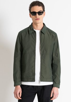 Chaqueta ligera verde con cremallera frontal, cuello y cinturilla elástica. Llevada sobre una camiseta blanca, combinada con pantalones negros y gafas de sol.
