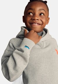 Sudadera gris de tejido suave, con un bolsillo canguro, puños acanalados y un logo azul en la manga, además de un diseño naranja en la parte frontal.